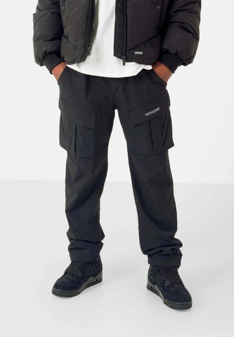 Schwarze Cargo-Hosen mit mehreren Fronttaschen, aus strapazierfähigem Material. Kombiniert mit einer schwarzen Pufferjacke und schwarzen Sneakers.