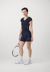 Top atlético azul marino con acentos blancos, combinado con pantalones cortos azul marino. El sujeto sostiene una raqueta de tenis y lleva zapatillas blancas y calcetines cortos.