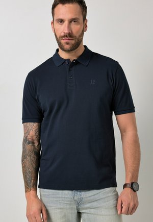 Mann mit Bart, trägt schwarzes Poloshirt und helle Jeans, zeigt detaillierten Tattoo-Ärmel am linken Arm und Armbanduhr am rechten Handgelenk.