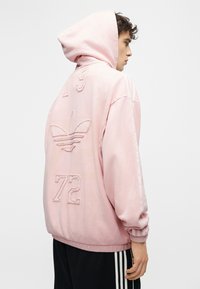 Personne portant un sweat à capuche rose avec le logo Adidas et les chiffres 19 et 72 au dos, associée à un pantalon noir Adidas avec des bandes blanches.