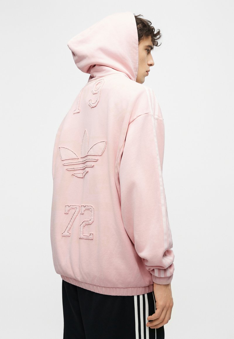 Personne portant un sweat à capuche rose avec le logo Adidas et les chiffres 19 et 72 au dos, associée à un pantalon noir Adidas avec des bandes blanches.