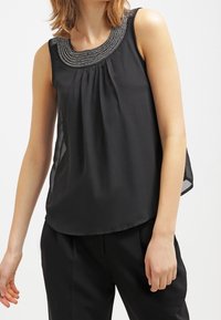 Top senza maniche nero con un frontale plissè e collo decorato con perline. Tessuto leggero e morbido, indossato con pantaloni neri.