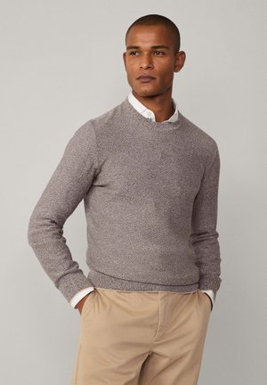 Pullover aus leicht braunem strukturiertem Strick, Rundhalsausschnitt, enganliegende Ärmel. Kombiniert mit einem weißen Hemd darunter und beigen Hosen.