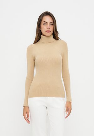Femme portant un pull à col roulé côtelé beige et un pantalon blanc, debout devant un fond clair uni, regardant droit devant elle.