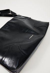 Borsa crossbody in pelle nera lucida con tracolla regolabile e piccolo logo argento su uno sfondo chiaro e semplice.