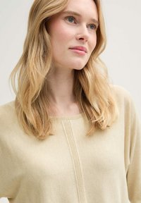 Beige, leichtes Strickoberteil mit weit ausgeschnittenem Hals und dezentem vertikalen Nahtdetail in der Mitte vorne. Weiche Textur.