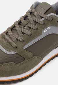 Tenis verde-oliva feitos de camurça e malha, com detalhes cinzentos, atacadores texturizados, uma entressola branca e uma sola preta com detalhes laranja.