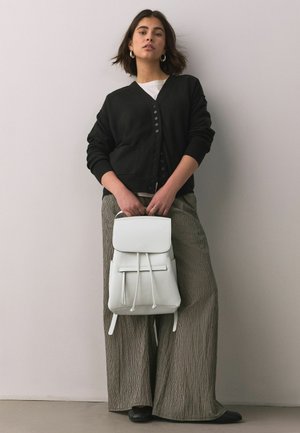 Jauna moteris dėvi juodą megztinį ir languotus plačiąsias kelnes, laiko baltą kuprinę pilkoje sienos fone.