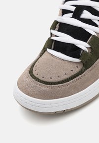 Beige ja oliivivihreät mokkanahka sneakerit, joissa on valkoiset nauhat ja teksturoitu varvaskuppi. Yksityiskohtina on rei'ityksiä ja kontrastiväriset ompeleet.