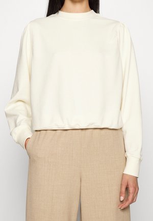 Femme portant un chemisier crème à manches longues avec un ourlet froncé et un pantalon beige taille haute à jambes larges, debout avec une main dans la poche.