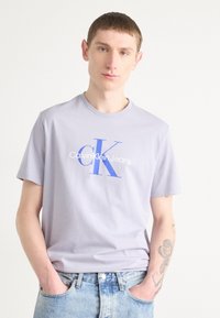 Light purple cotton T-shirt featuring a blue "CK" logo and "Calvin Klein Jeans" text. Short sleeves and crew neckline, paired with blue jeans.
