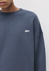 Tengerészkék pulóver bordás kerek nyakkal és hosszú ujjakkal. A bal mellrészen egy kicsi, fehér hímzett logó található. Puha, texturált anyag.