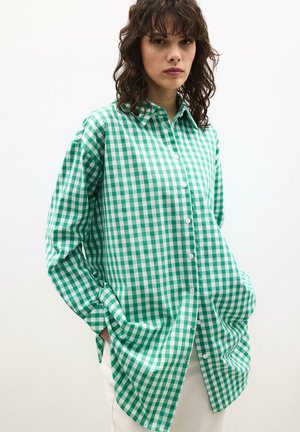 Femme portant une chemise à boutons surdimensionnée à carreaux verts et blancs, les mains dans les poches, sur un fond uni.
