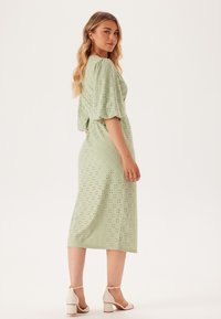 Abito midi verde con dettagli in pizzo, maniche puff e vita aderente. Presenta uno spacco sul retro ed è abbinato a sandali con tacco quadrato di colore chiaro.