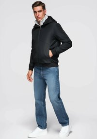 Młody mężczyzna stoi z jedną ręką w kieszeni, ubrany w czarną kurtkę na zamek, niebieskie dżinsy i białe trampki na jednolitym tle.