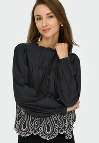 Blouse noire à manches longues, avec un col froncé et une broderie décorative blanche formant un motif de paisley à l'ourlet.