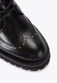 Scarpa in pelle nera con finitura lucida, caratterizzata da borchie decorative dorate e una punta lavorata, montata su una suola in gomma nera spessa.