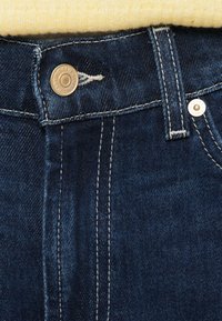 Temno modre kavbojke iz jeansa z robustnim pasom, kovinskim gumbom na vrhu, kontrastnim belim šivom in gladko teksturo.