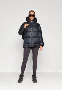 Frau in einer schwarzen Daunenjacke, schwarzen Leggings, Wanderschuhen, Sonnenbrille und mit einem schwarzen Rucksack vor einem einfarbigen hellen Hintergrund.