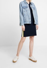 Jasnoniebieska kurtka dżinsowa, dopasowana, z kieszeniami na piersi. Czarna sukienka z żółtym paskiem po bokach, białe sneakersy z plecioną podeszwą.