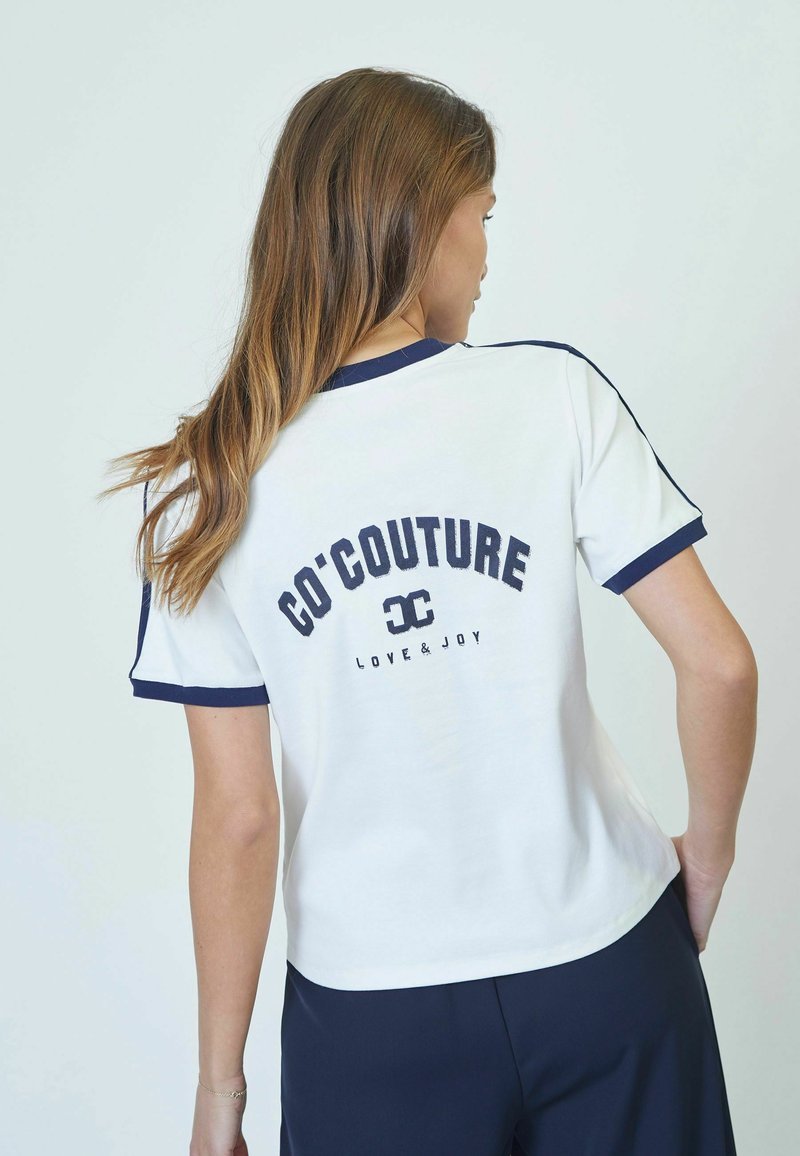 Camiseta blanca con ribete marino, que presenta "CO'COUTURE" y "LOVE & JOY" impresos en la parte trasera. Manga corta, corte relajado. Textura de algodón suave.