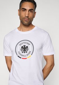 Fehér pamut póló, amelyen kör alakú fekete, szürke és piros grafikus logó található, rajta egy sas és a "DEUTSCHER FUSSBALL-BUND" felirat. Rövid ujjú.