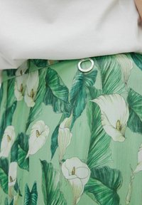Grøn nederdel med et botanisk print af hvide calla-liljer og grønne blade, med et sølvfarvet ring-akkent. Glat, struktureret stof.