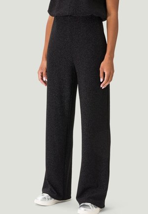 Osoba nosząca czarne spodnie z wysokim stanem i szerokimi nogawkami, w połączeniu z srebrnymi tenisówkami, stojąca z luźno opuszczonymi rękami po bokach.