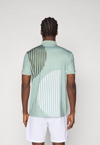 Polo a maniche corte in verde menta con motivi geometrici, caratterizzato da strisce verticali e forme circolari in verde scuro e bianco.