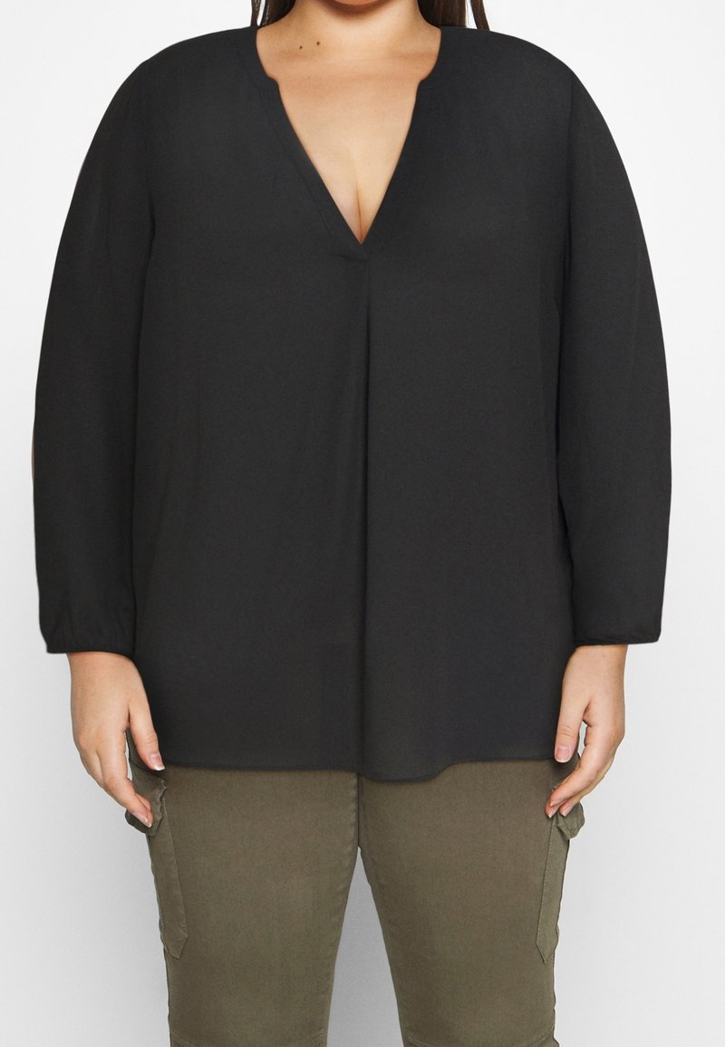 Blusa nera a maniche lunghe con scollatura a V, realizzata in tessuto morbido. Presenta un fit rilassato e cuciture minime. Abbinata a pantaloni cargo verde oliva.