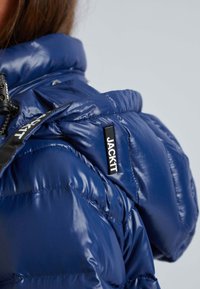 Personne portant une doudoune bleue brillante avec une étiquette de marque "JACKIT" en noir et blanc visible sur la zone de l'épaule.