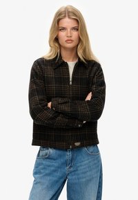 Zwarte en bruine geruite ritsjack, gemaakt van textuurstof, met een kraag en verstelbare mouwen. Gecombineerd met lichtblauwe spijkerbroek.