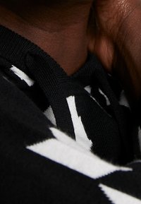 Gros plan sur un cou à peau foncée portant un vêtement en maille noire avec des rayures blanches audacieuses verticales et diagonales.