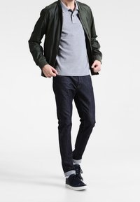Pánský outfit s tmavě zelenou bomber bundou, šedým polo tričkem, tmavými džínami a navy teniskami s bílou podrážkou.