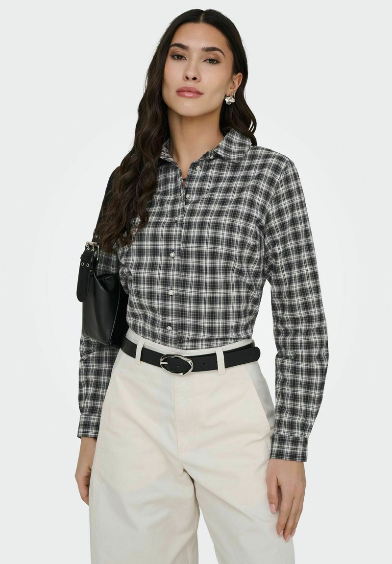 Femme aux longs cheveux foncés portant une chemise à carreaux noir et blanc boutonnée, un pantalon blanc avec une ceinture noire, et portant un sac noir à l'épaule.
