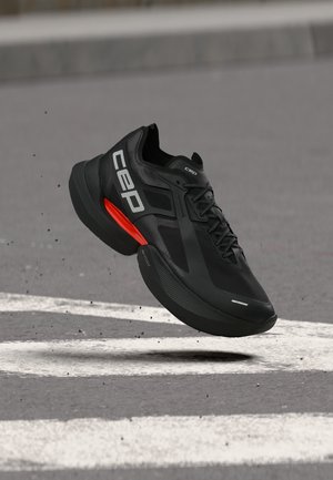 Black athletic shoe with white CEP logo and orange midsole detail hovering above an asphalt surface with white road markings.