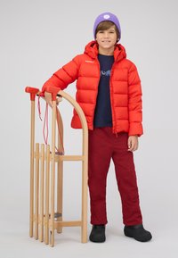 Rode pufferjack, marineblauwe shirt, rode broek en zwarte laarzen. Houten slee met rode accenten ernaast gehouden. Paarse beanie met witte ronde patch.