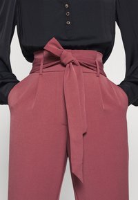 Hoogwaardige roze broeken met een stoffen riem die in een strik is geknoopt, met zijzakken en een gladde textuur. Zwarte blouse met knoopdetails.