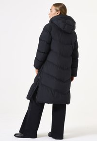 Manteau en nylon noir avec capuche, présentant des coutures en chevrons, des poches zippées sur les côtés et une coupe mi-longue. Porté sur un pantalon sombre à jambes larges.