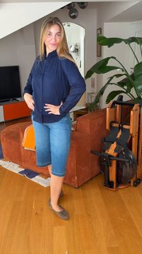 Νεαρή γυναίκα με μπλε πουκάμισο και τζιν σορτς στέκεται στο σαλόνι με πορτοκαλί καναπέ, μεγάλο πράσινο φυτό και ξύλινο μηχάνημα γυμναστικής.