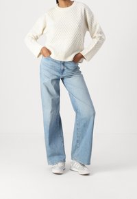 Weißer Strickpullover mit strukturiertem Muster, kombiniert mit hellblauen weit geschnittenen Jeans und grauen Sneakern mit weißen Akzenten.