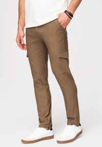 Homme portant un pantalon cargo marron slim-fit avec poches latérales à rabat, assorti à des baskets blanches et une chemise blanche, debout devant un fond uni.