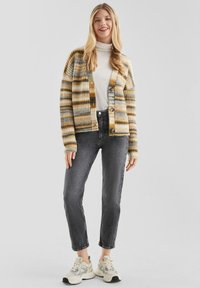 Gestreifte Strickjacke in Gelb, Grau und Beige, mit Knopfverschluss, lockerer Passform und geripptem Material. Getragen mit grauen Jeans und Sneakers.