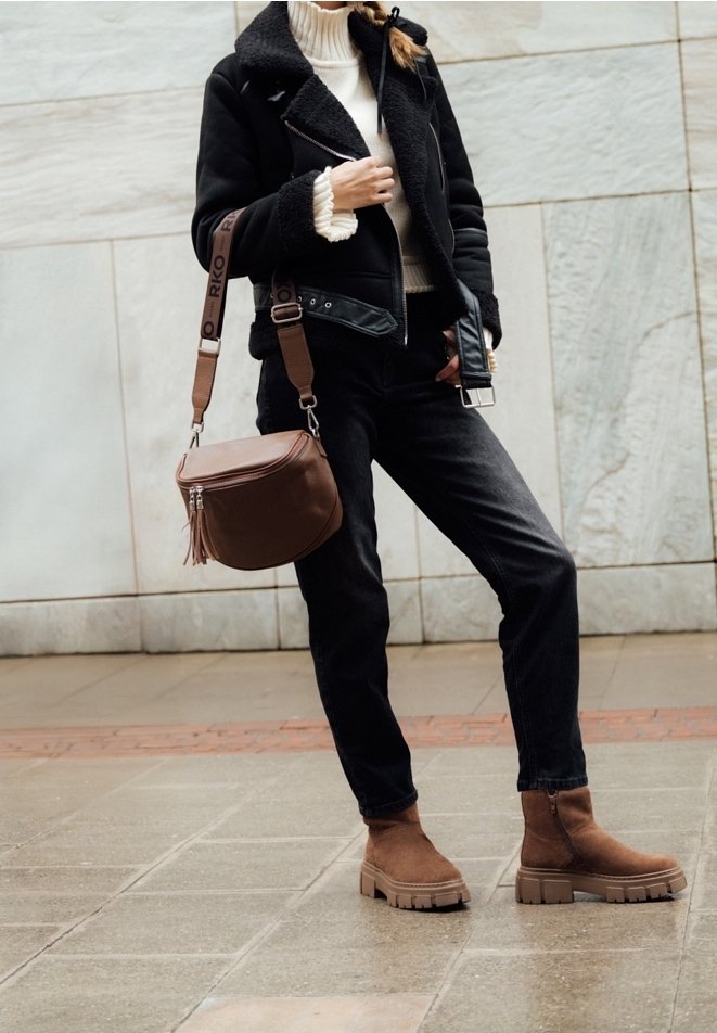 Femme portant une veste en peau lainée noire, un col roulé blanc, un jean noir, des bottes marron, et portant un sac bandoulière en cuir marron, debout à l'extérieur.