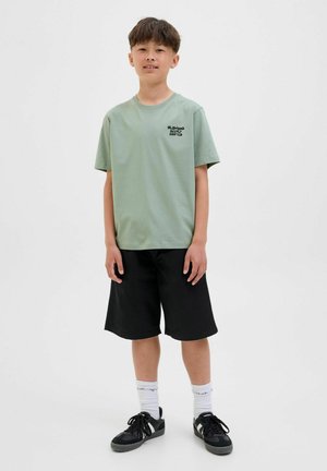 Lichtgroen katoenen T-shirt met korte mouwen en zwarte tekst, gecombineerd met zwarte knielange shorts en zwarte sneakers met witte sokken.