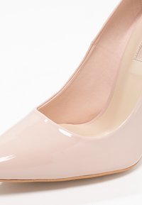 Ljust rosa lackläders högklackad sko med spetsig tå och slät yta. Interiören har en beige foder; minimalistisk design.