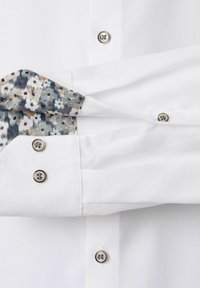 Weißes, strukturiertes Hemd mit silbernen Knöpfen und floral gemusterter Innenmanschette, die teilweise umgeschlagen ist.