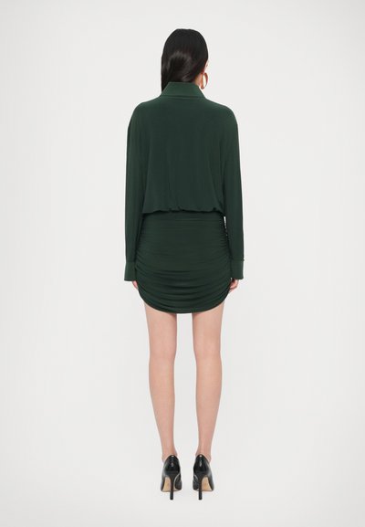 Tmavozelené šaty s dlhým rukávom, golierom a priliehavou, žabkovanou sukňou. Jemná látka a priliehavý strih, doplnené čiernymi lodičkami.