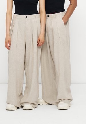 To personer iført beige bukser med vide ben, sorte toppe og hvide sneakers, står side om side mod en hvid baggrund.
