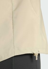 Hellbeige Jacke mit glatter Textur, Kordelzug am Saum und seitlichen Taschen. Teilweise vor einem dunklen Material gezeigt.